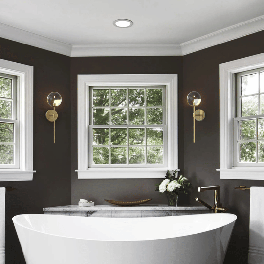 Elegant bathroom with a white freestanding bathtub positioned beneath three large windows framed in white trim. The walls are painted dark gray, accented with modern brass wall sconces featuring globe lights. A gold faucet and a vase of white flowers on the tub ledge add a touch of sophistication.