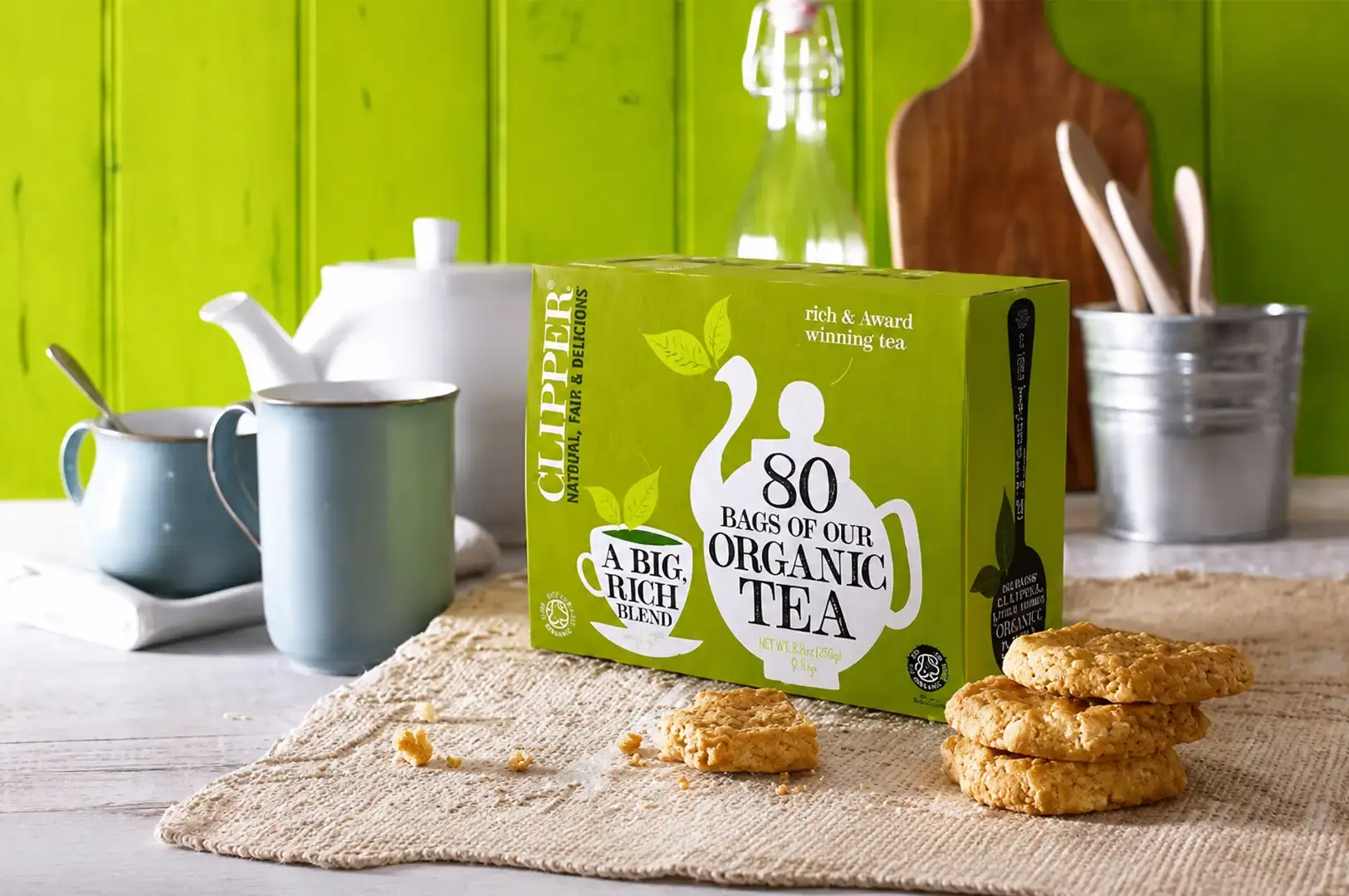 A bright, rustic kitchen scene displaying a green Clipper box labeled "80 Bags of Our Organic Tea" resting on a woven burlap placemat. To the right of the tea box sits a stack of golden oat cookies with scattered crumbs, while a white ceramic teapot, a light blue mug, and a vintage glass bottle are subtly arranged in the background against a vibrant lime-green wooden panel wall.