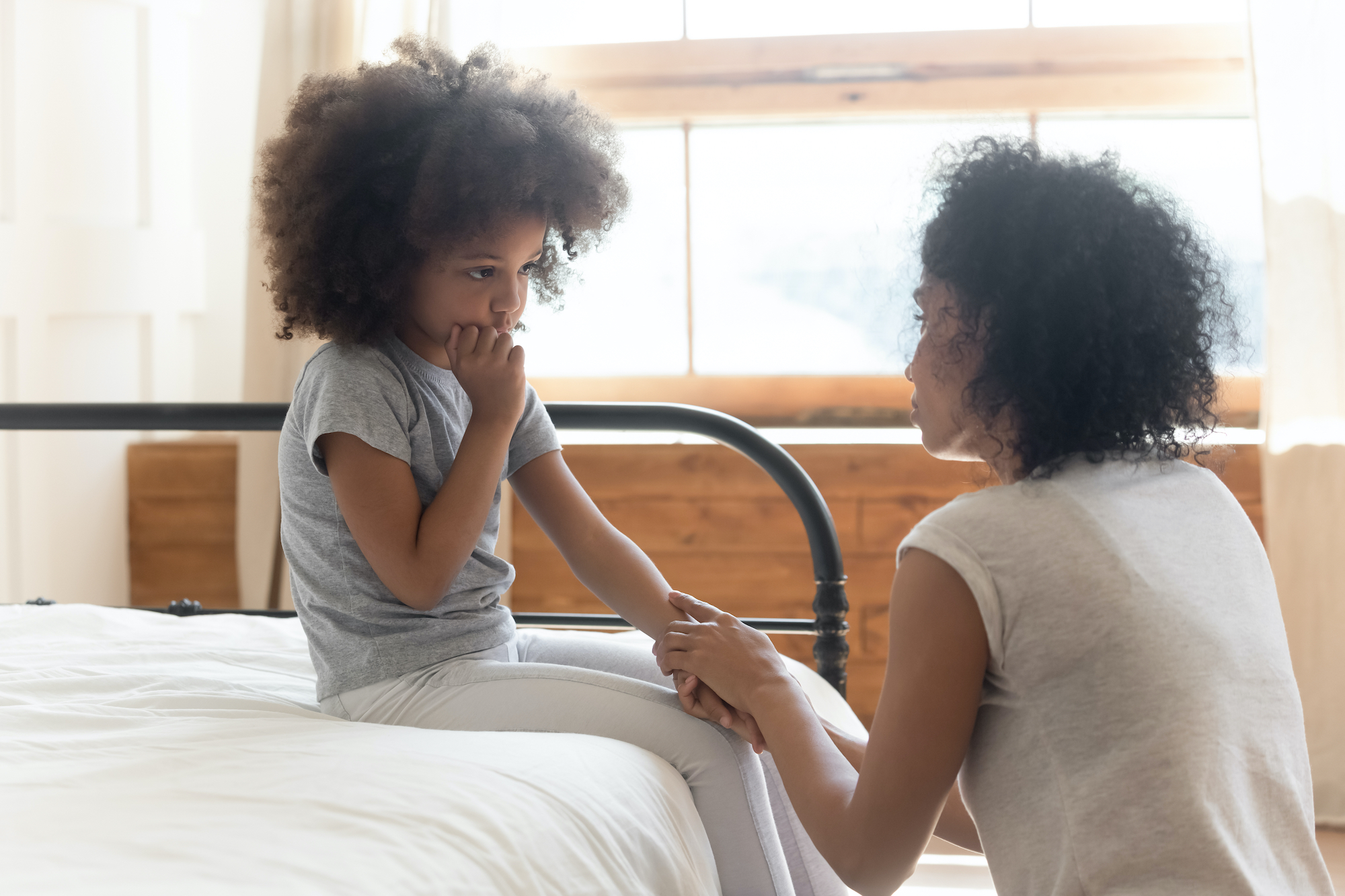 Little black girl sitting on a bed looking unsure and her mother is kneeling in front of her comforting her