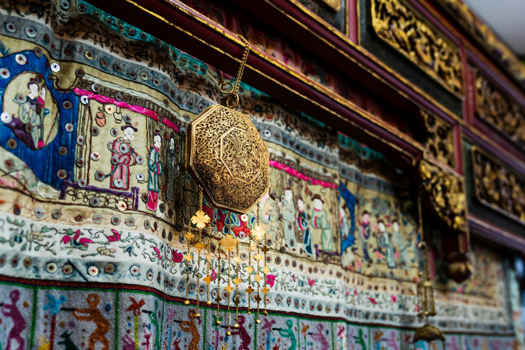  An ornate, golden octagonal pendant with dangling floral charms hangs against a highly detailed tapestry featuring embroidered figures and sequins. The background includes dark, intricately carved wooden panels, suggesting a setting rich in traditional craftsmanship and cultural heritage.