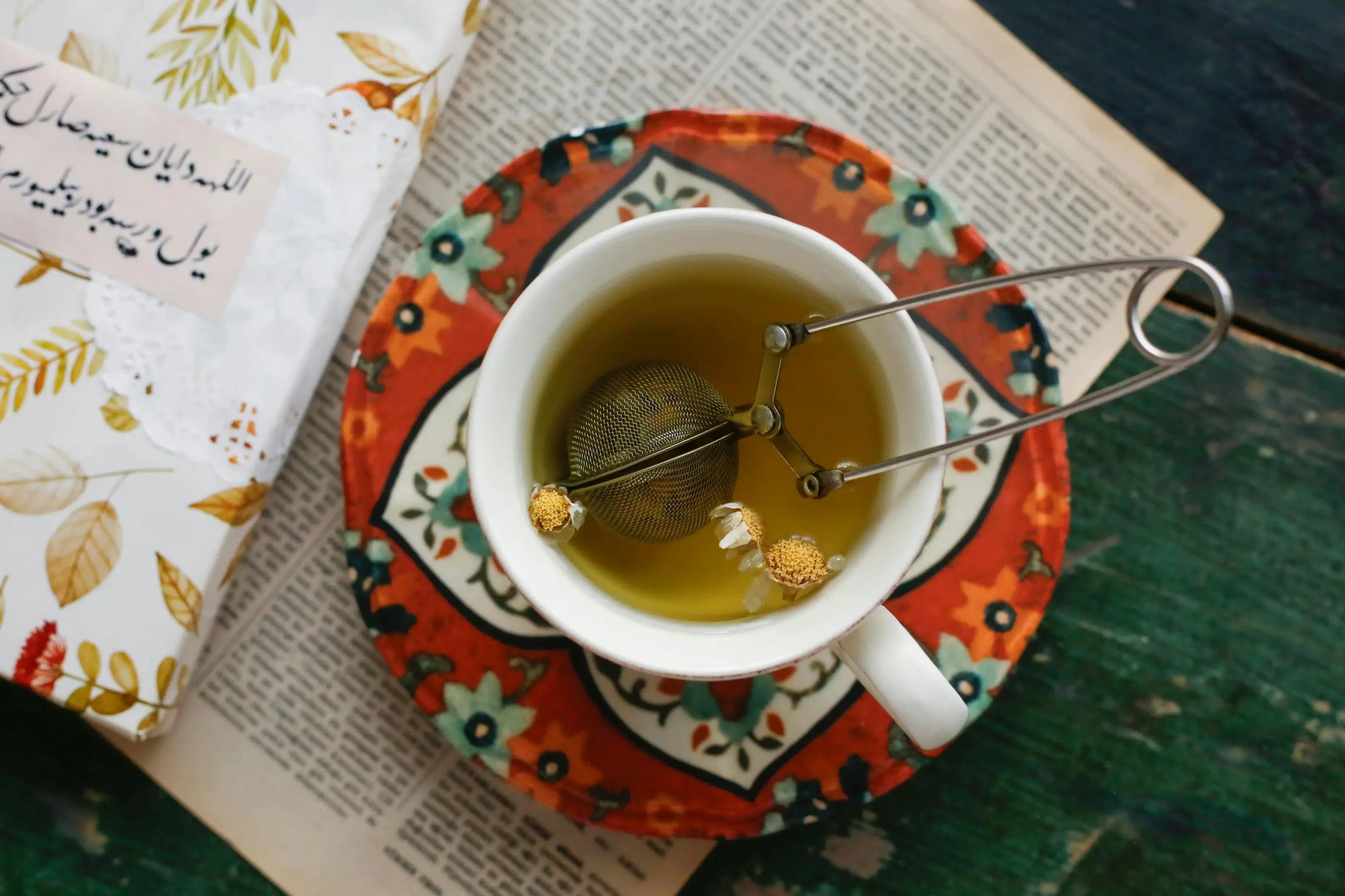 An overhead view shows a cup of chamomile tea containing a metal mesh infuser spoon and loose flowers, resting on a colorful, patterned saucer. The tea is set against a background of an open newspaper and a book with Arabic script on its cover.