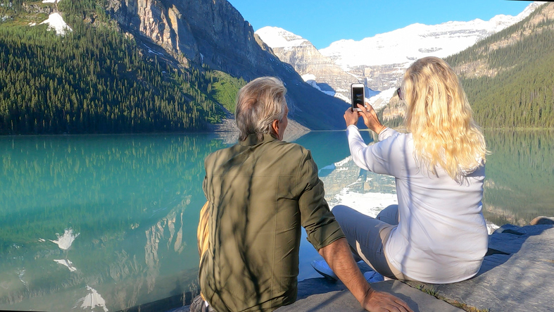 Gray-haired man and blonde woman sitting on a rock and taking a photo of mountains. 