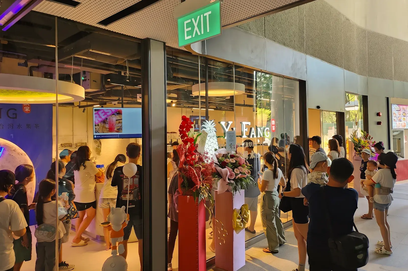 Bustling storefront and queue of customers outside a Yi Fang Taiwan Fruit Tea shop adorned with floral grand opening stands.