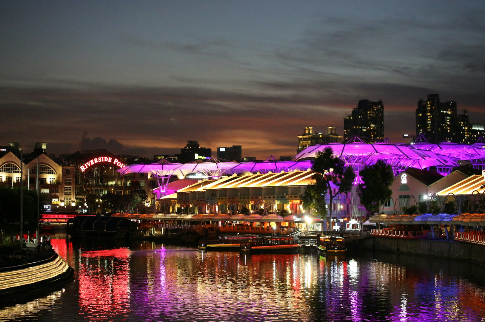Night view of a lively riverside area with glowing purple and yellow lights reflecting on the water. The sky is dusky, and buildings are in the background.