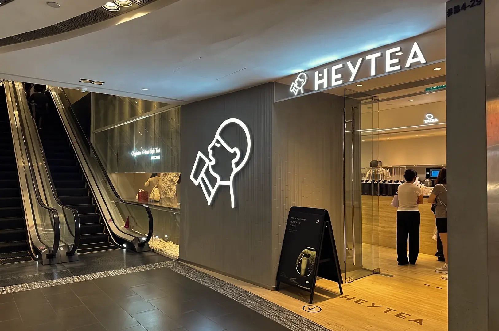 Storefront of a HEYTEA shop featuring a minimalist design, glowing logo, and customers standing at the service counter next to an escalator.
