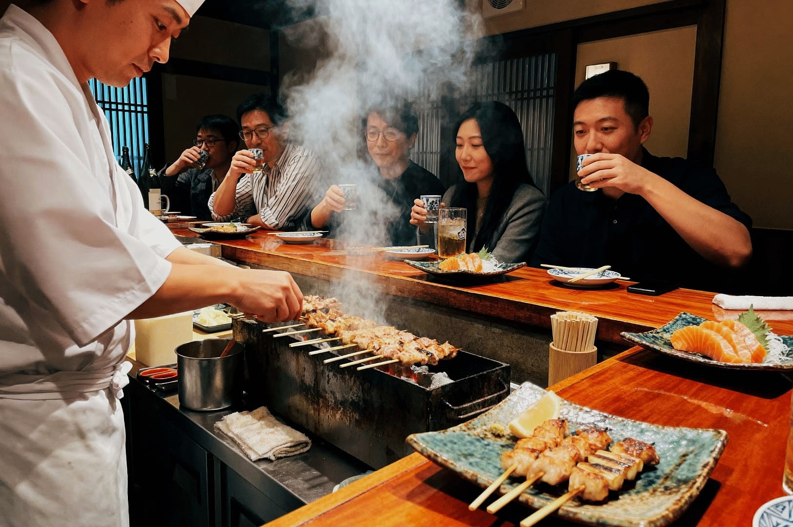 A chef in a white uniform grills skewers at a smoky bar, while diners seated at a wooden counter enjoy drinks. Plates of sushi and skewers are served. The atmosphere is warm and inviting.