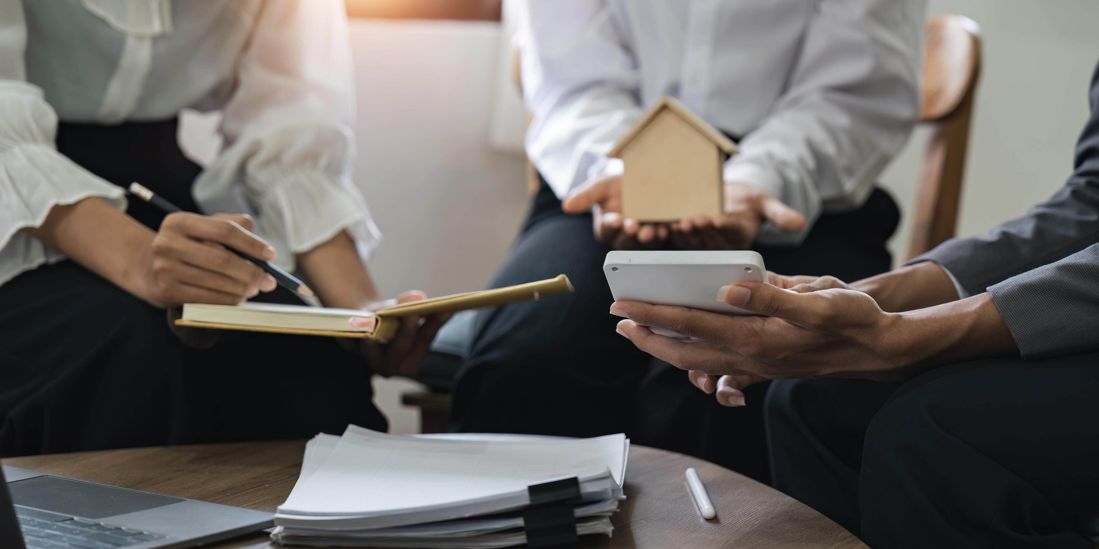 Three people are in a meeting with documents and technology. One of the people holds a model house in their hands.