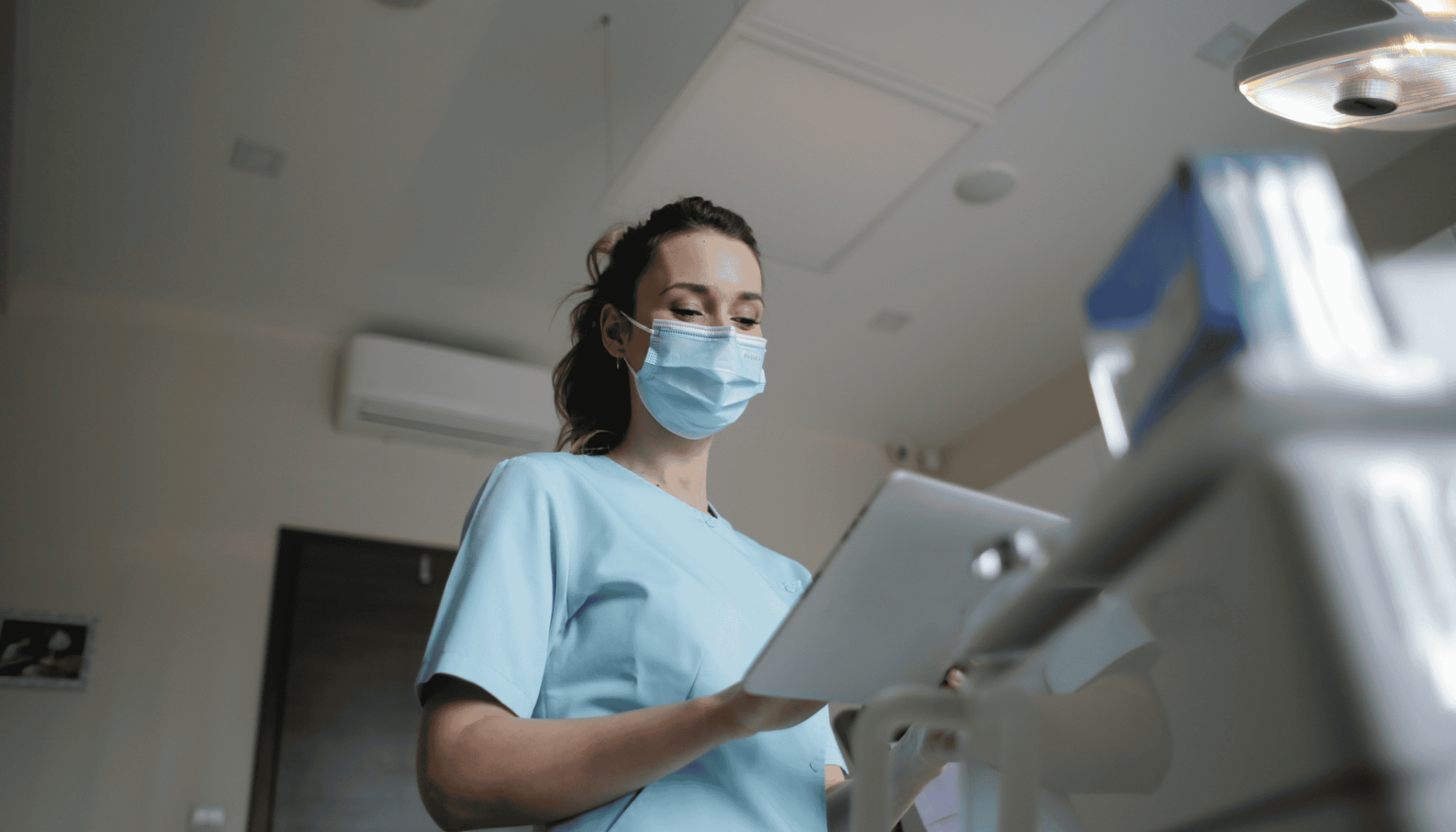 Kvinnelig tannhelsepersonell med munnbind og blå uniform ser på et nettbrett inne på en moderne tannklinikk.