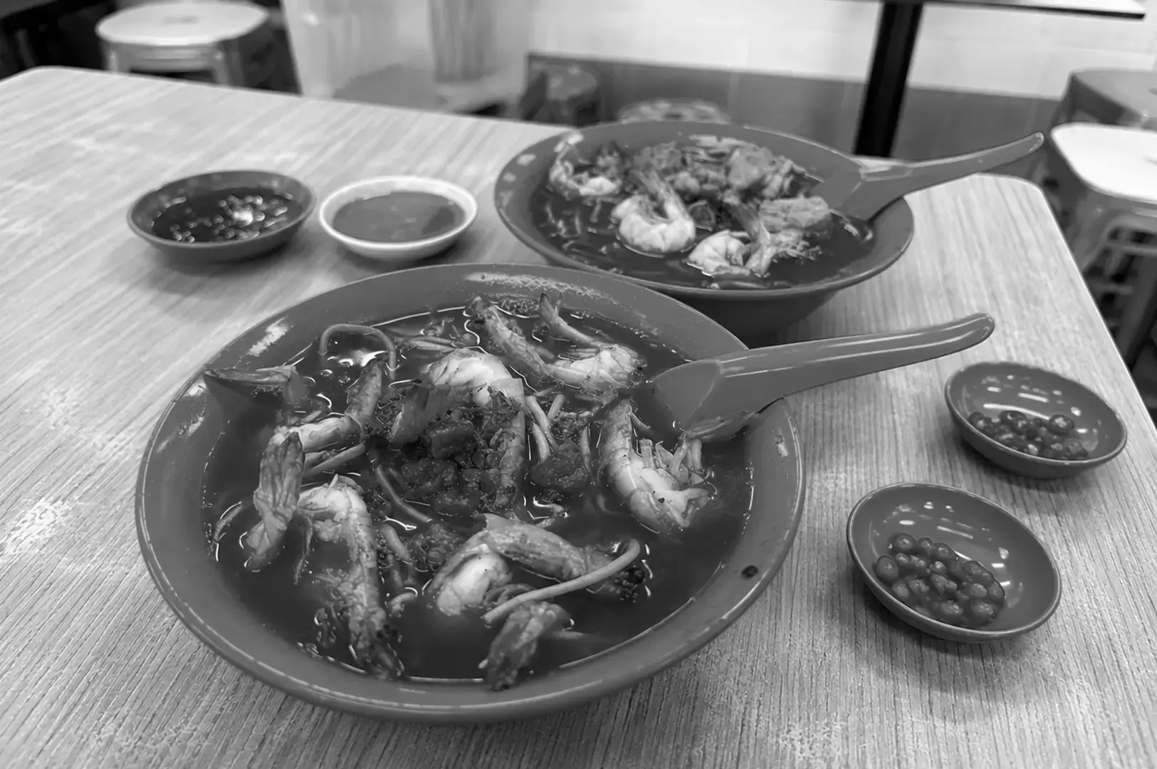 Two bowls of rich prawn noodle soup with dipping sauces on a wooden table.