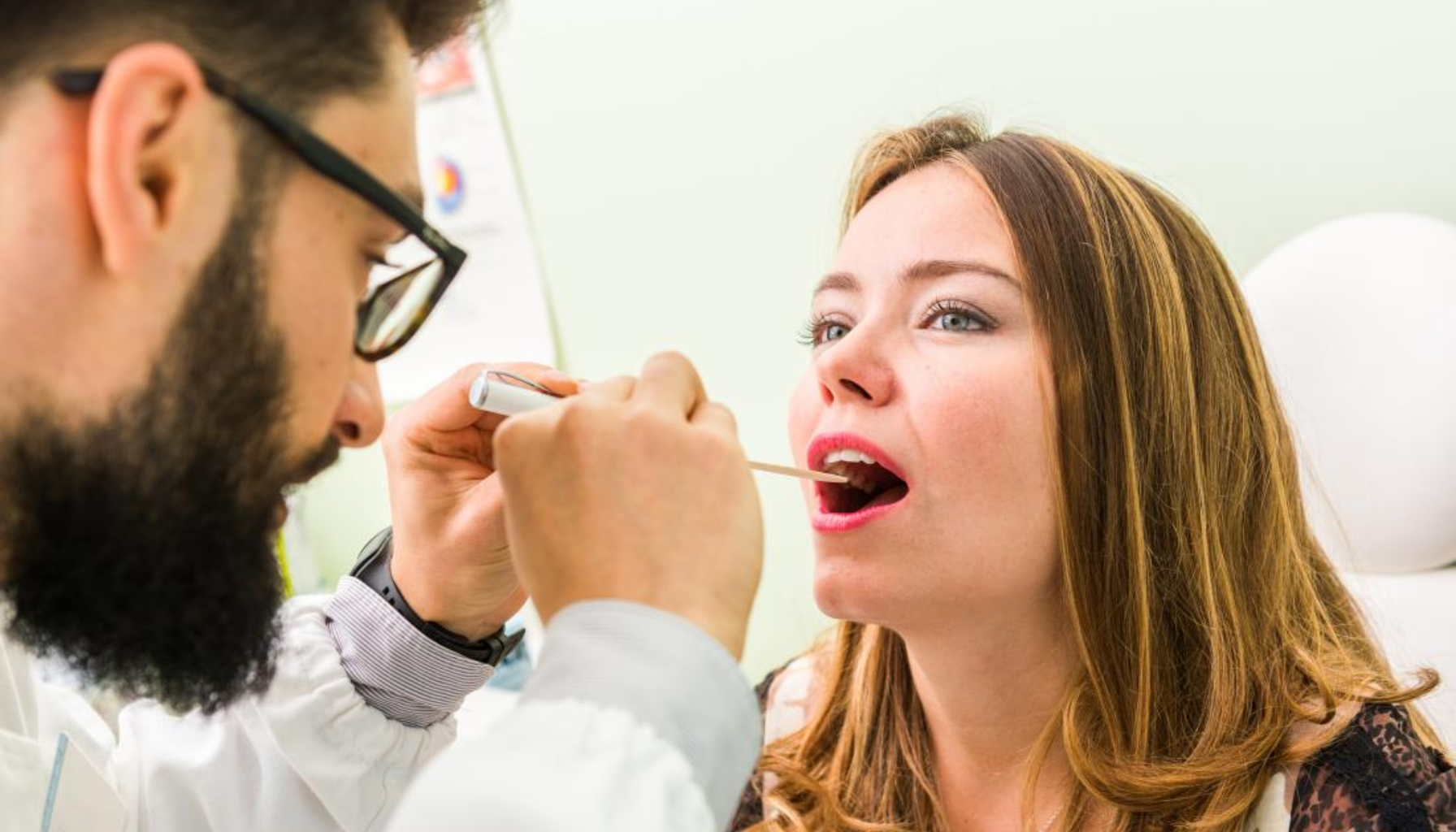 Grønn tunge – Hva det betyr og hvordan behandle det 19 "Lege undersøker en kvinnes hals med tungespatel under en medisinsk kontroll."