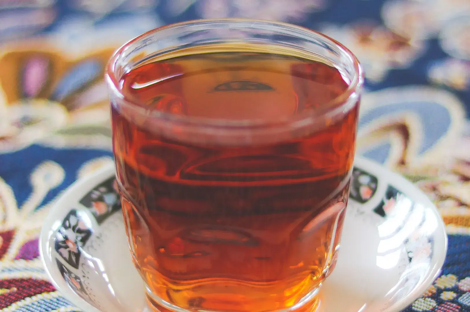 Close-up of a small glass cup filled with clear amber-red hot tea, resting on a patterned saucer over a colorful tablecloth.