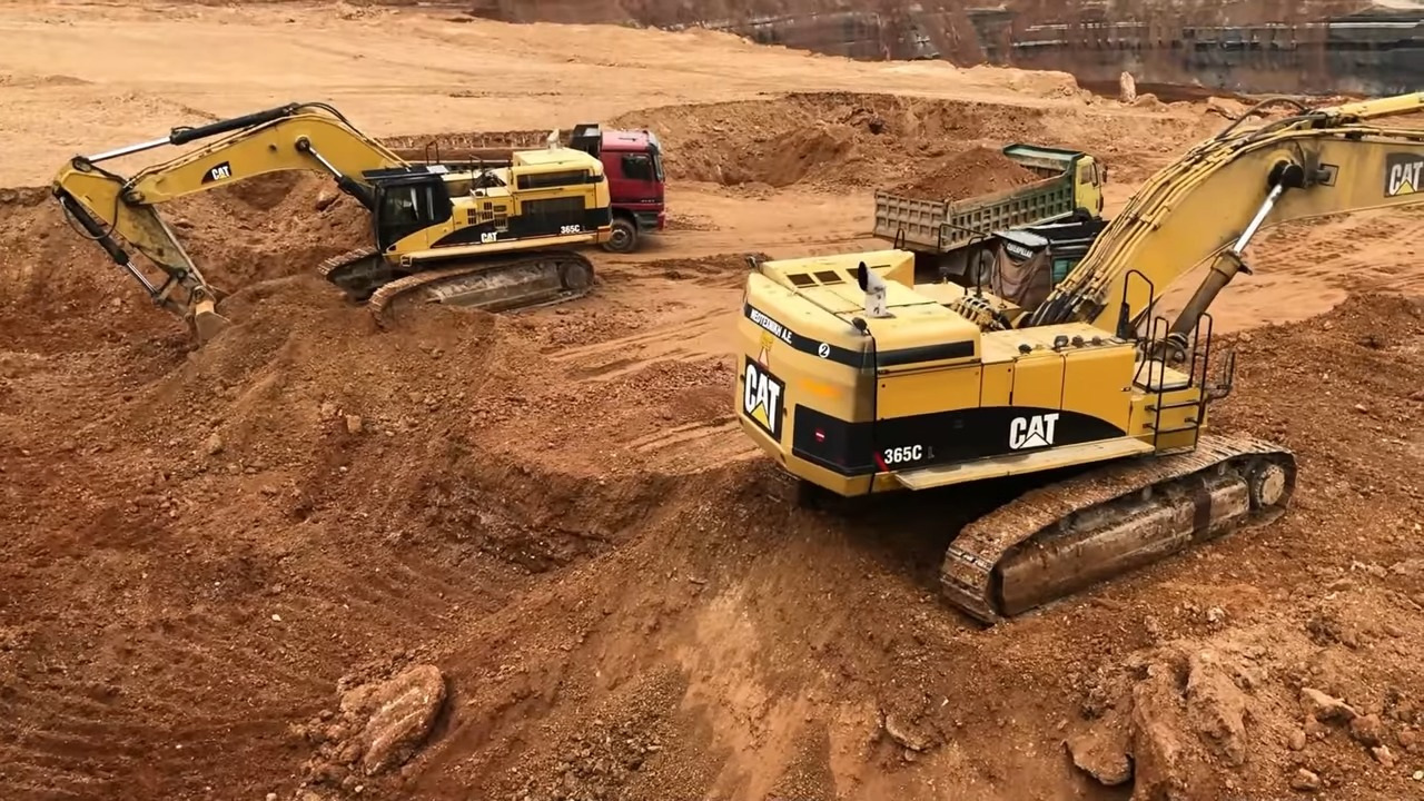Two yellow Caterpillar excavators digging and moving dirt in a large construction site with a red truck in the background