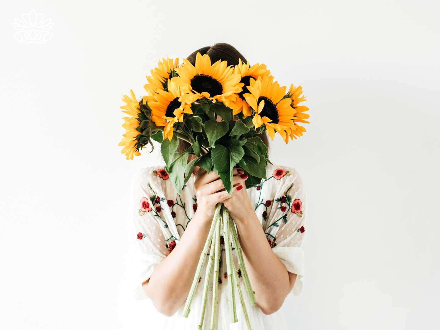A woman holding a large bouquet of vivid yellow sunflowers in front of her face, wearing a white floral dress, from the Sunflowers Collection by Fabulous Flowers and Gifts.