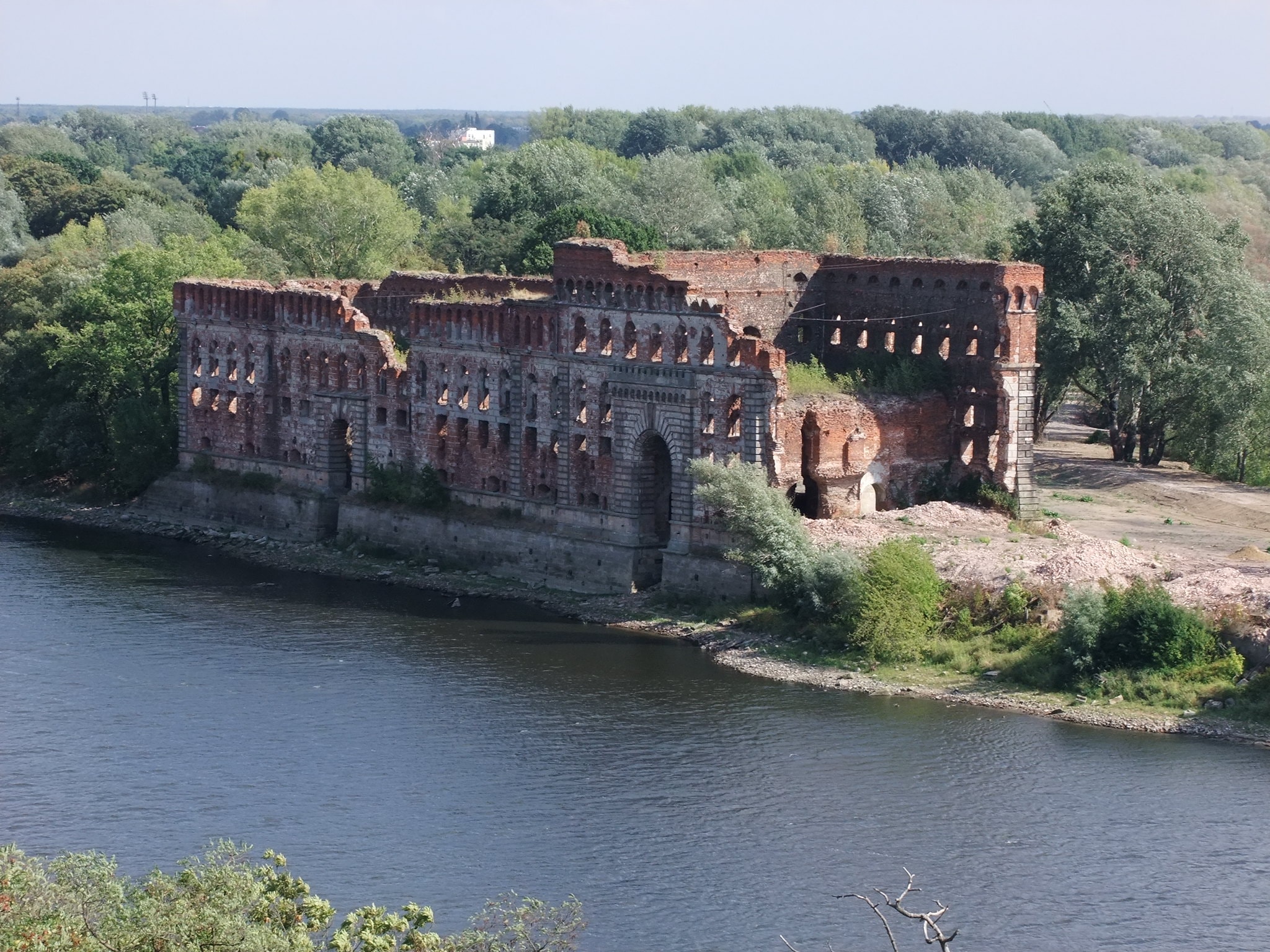 Ruiny fortu nad rzeką, z czerwonej cegły, otoczone zielenią i drzewami. Opuszczony, częściowo zawalony budynek, świadczący o minionej epoce i dawnej architekturze obronnej. (źródło: https://www.flickr.com/photos/maniak713/21144209888)