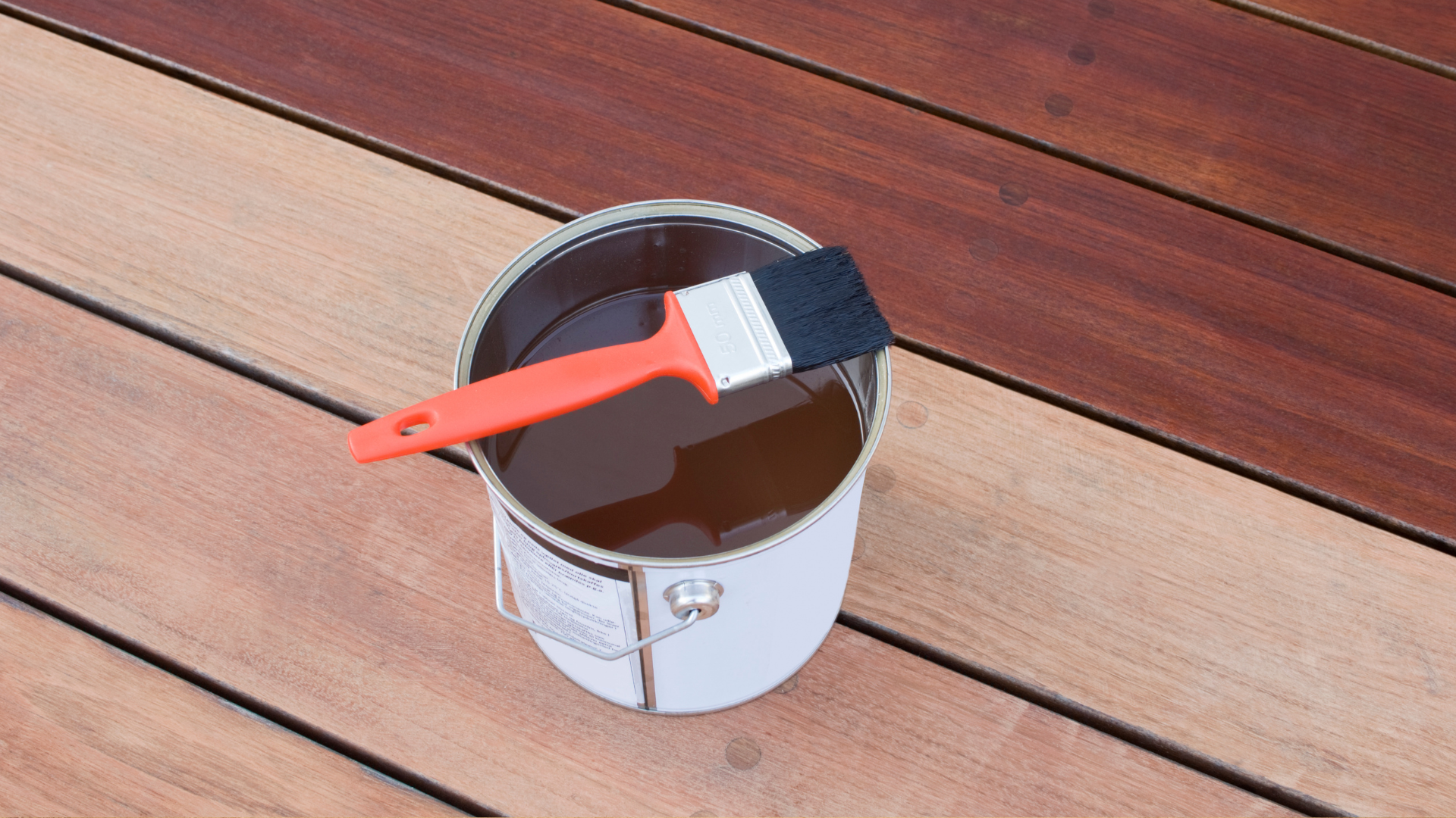 A can of brown wood stain on a deck, with some boards soaked in stain.