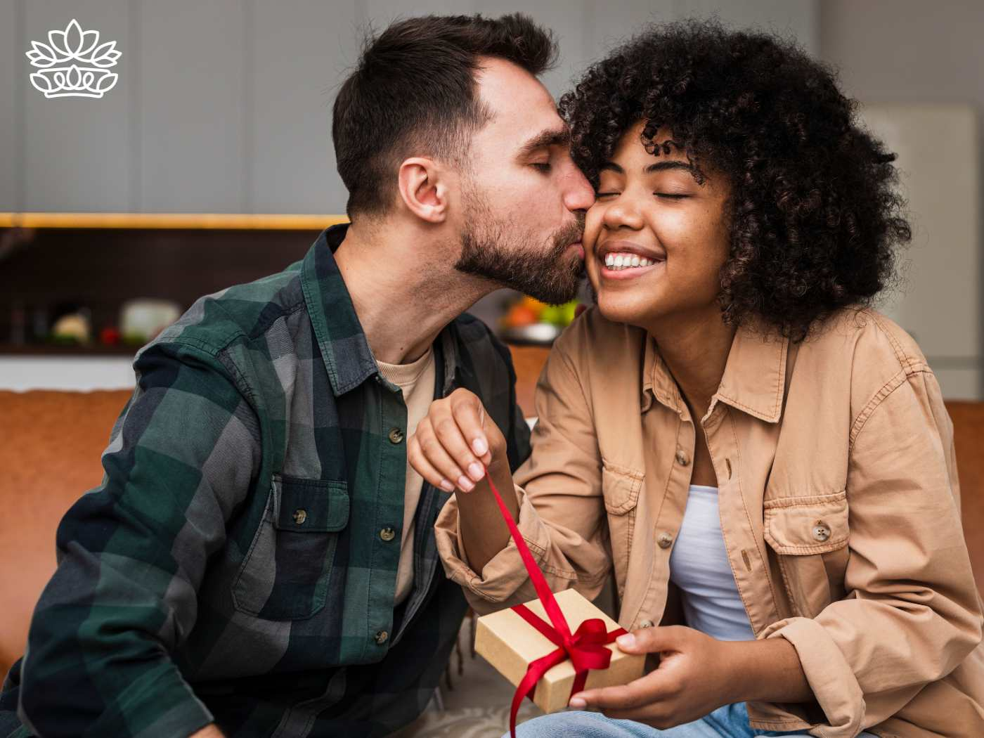 A man kissing a smiling woman on the cheek as she opens a gift box tied with a red ribbon, representing the Gift Boxes for Her Collection - Gift hampers and best gift ideas - Fabulous Flowers and Gifts