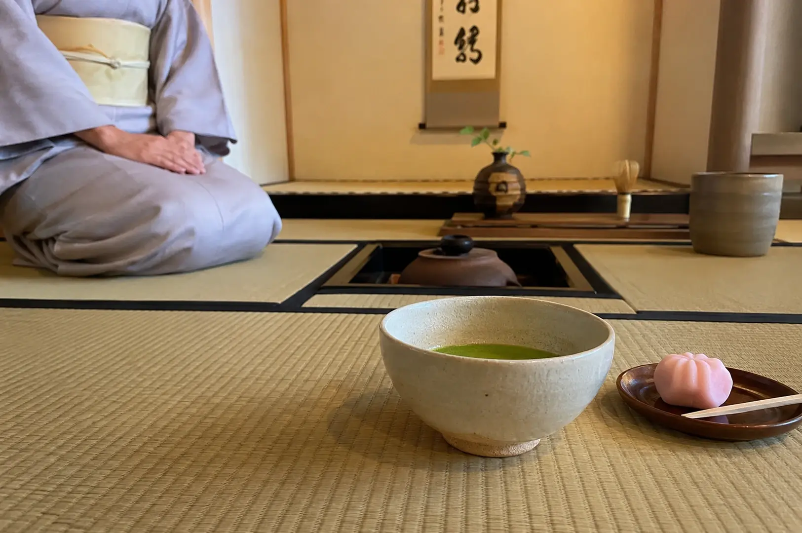 A traditional tea ceremony scene capturing a guest or host dressed in a light purple kimono kneeling in the formal seiza posture on a tatami mat. In the foreground, a wide, light-colored ceramic bowl holding prepared green matcha tea rests next to a small wooden plate containing a delicate, pink flower-shaped wagashi sweet with a small pick. In the middle ground, a sunken hearth holds a steaming iron kettle, while the background features a minimalist alcove with a hanging scroll and a simple ceramic vase holding a single green branch.