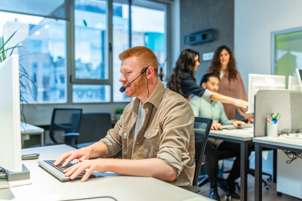 Customer support representative with a headset typing on a keyboard in a shared office space, showcasing effective business communication powered by upgraded business phone systems.