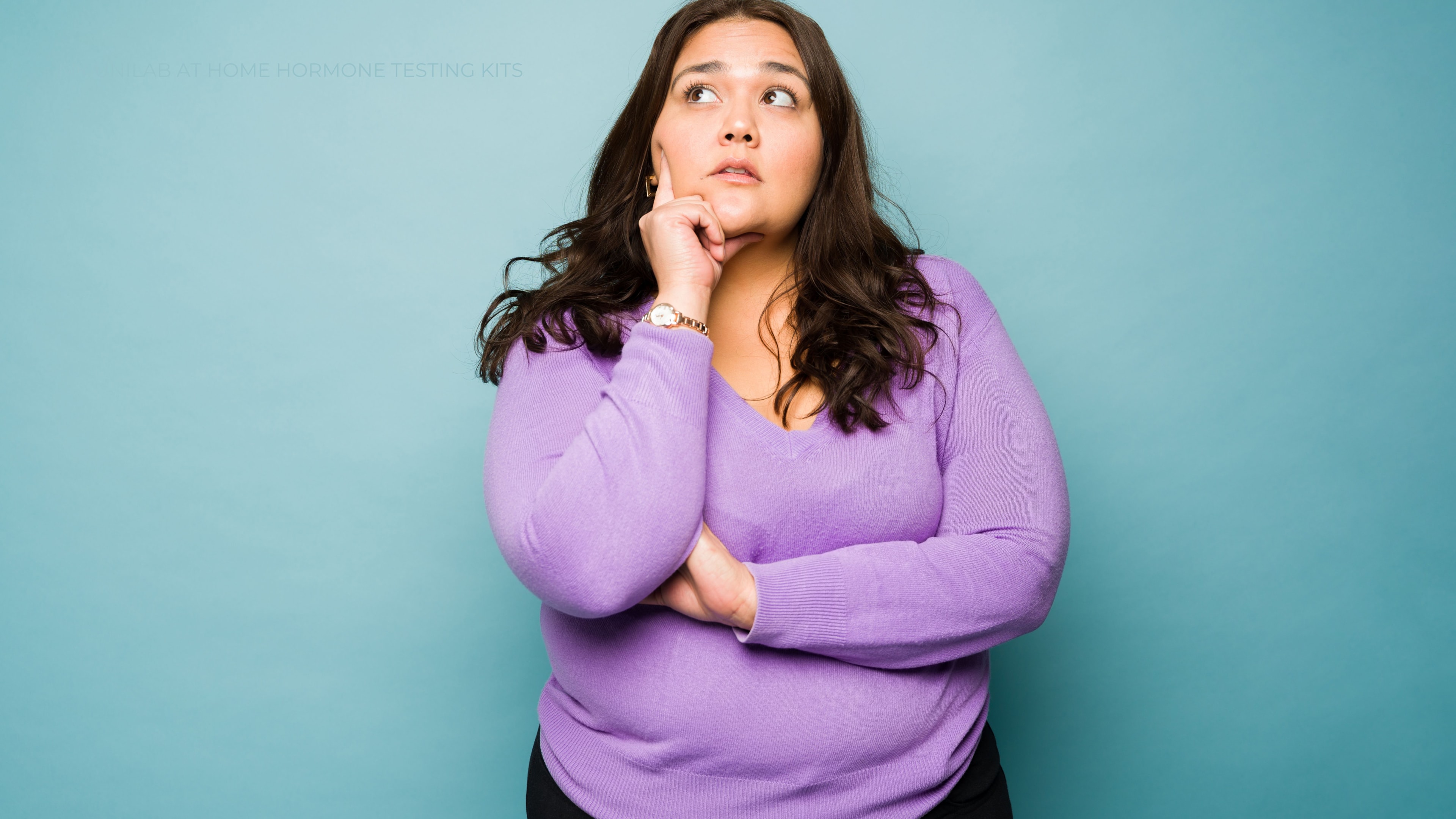 Women standing in front of a pale blue wall with her hand against on her face and the other arms crossed looking upward with a questioning expression.