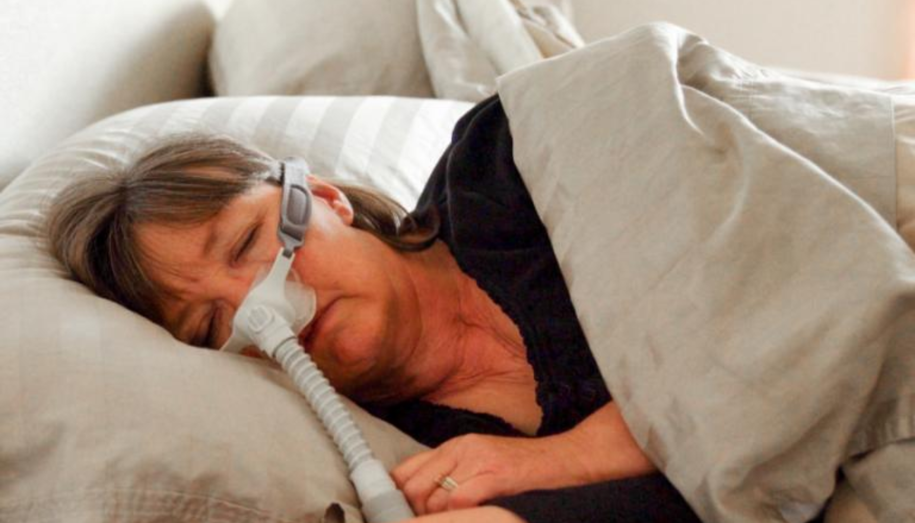 Eldre kvinne sover på siden med CPAP-maske og pusteslange, delvis dekket av dyne.