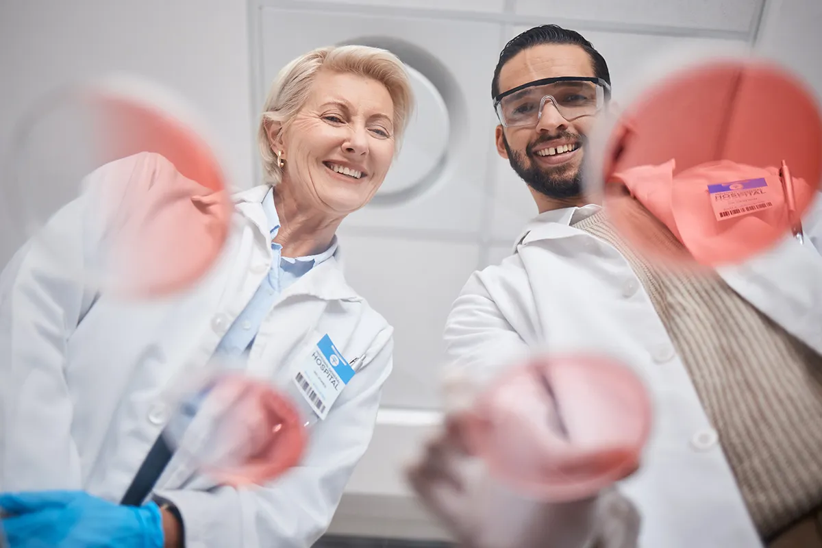 Dental professionals wearing white lab coats and protective eyewear smiling while holding dental equipment with pink protective barriers.