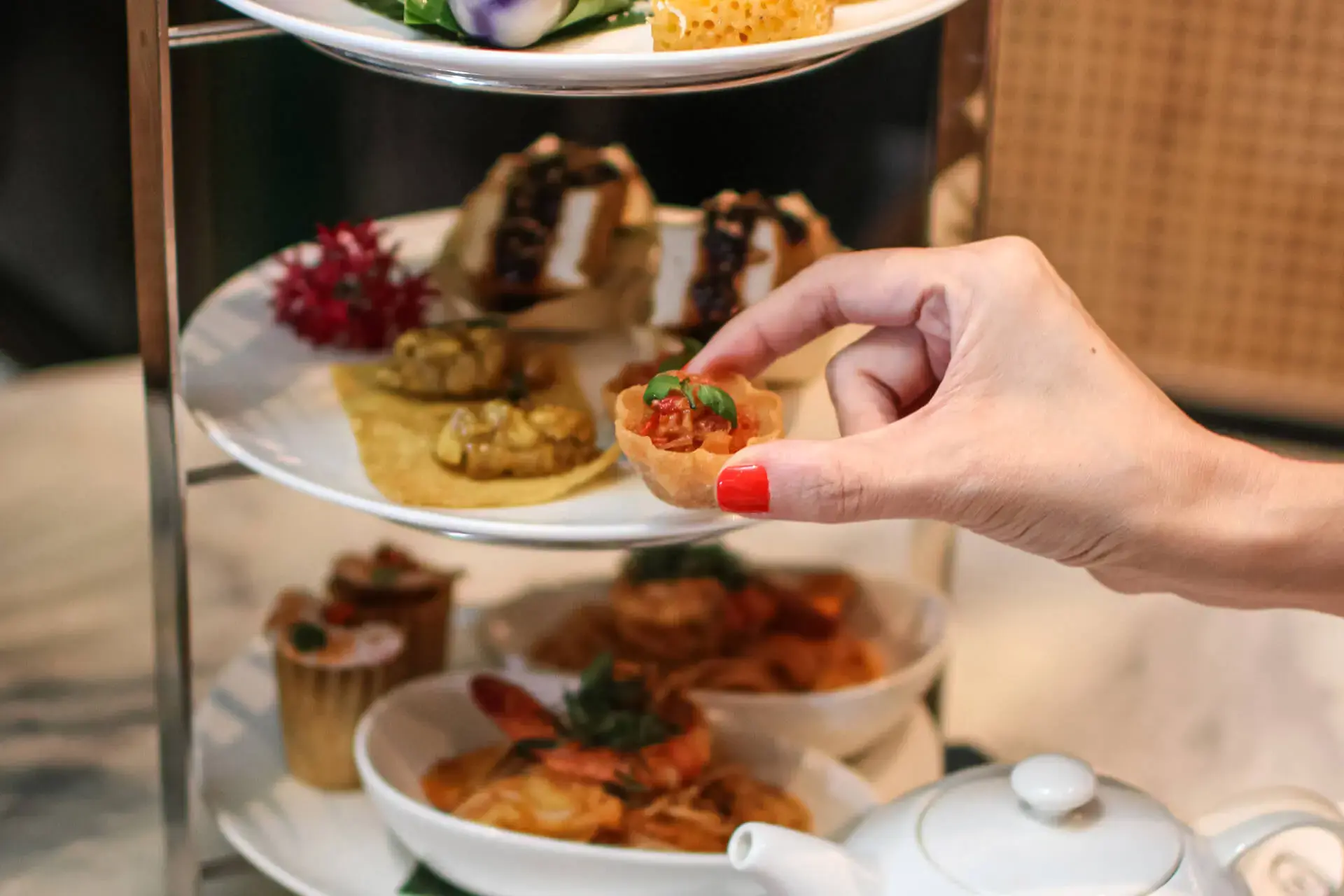A close-up shot shows a person's hand with red painted nails picking up a small pastry cup filled with a savory topping from a three-tiered high tea stand. The stand is filled with an assortment of Peranakan-style sweet and savory delicacies, accompanied by a white teapot in the foreground.