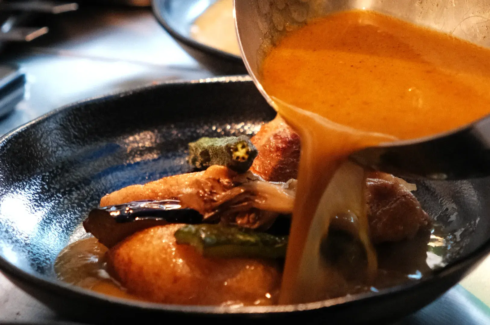 A dish being plated with a rich sauce being poured over vegetables and protein in a dark bowl.