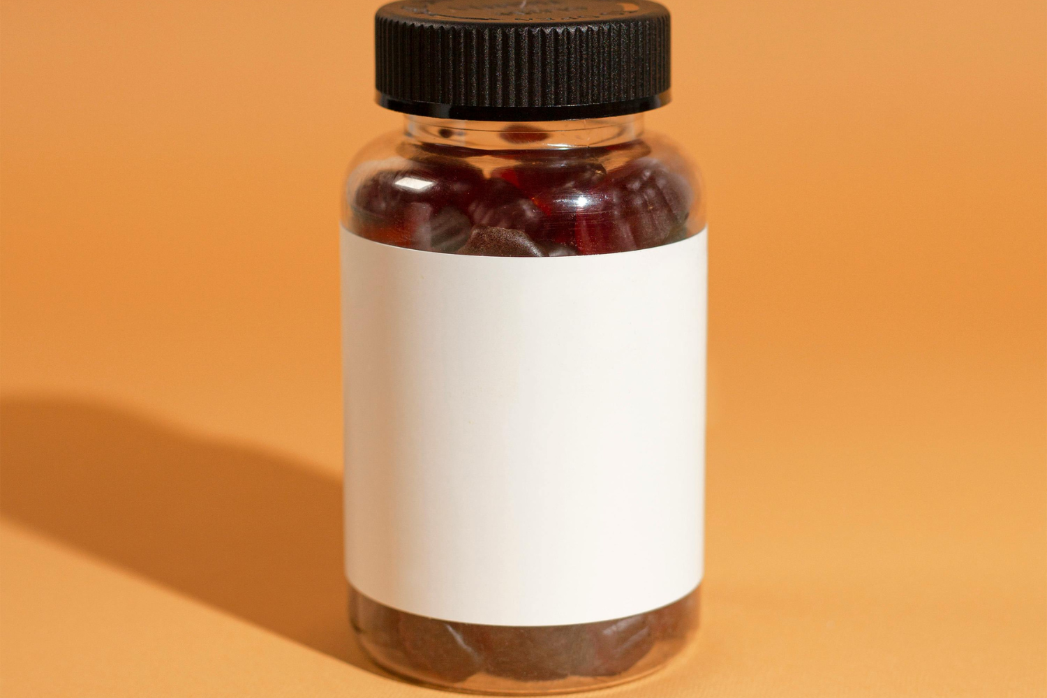 A bottle with a black cap and blank white label, filled with red capsules, sits on a peach background.
