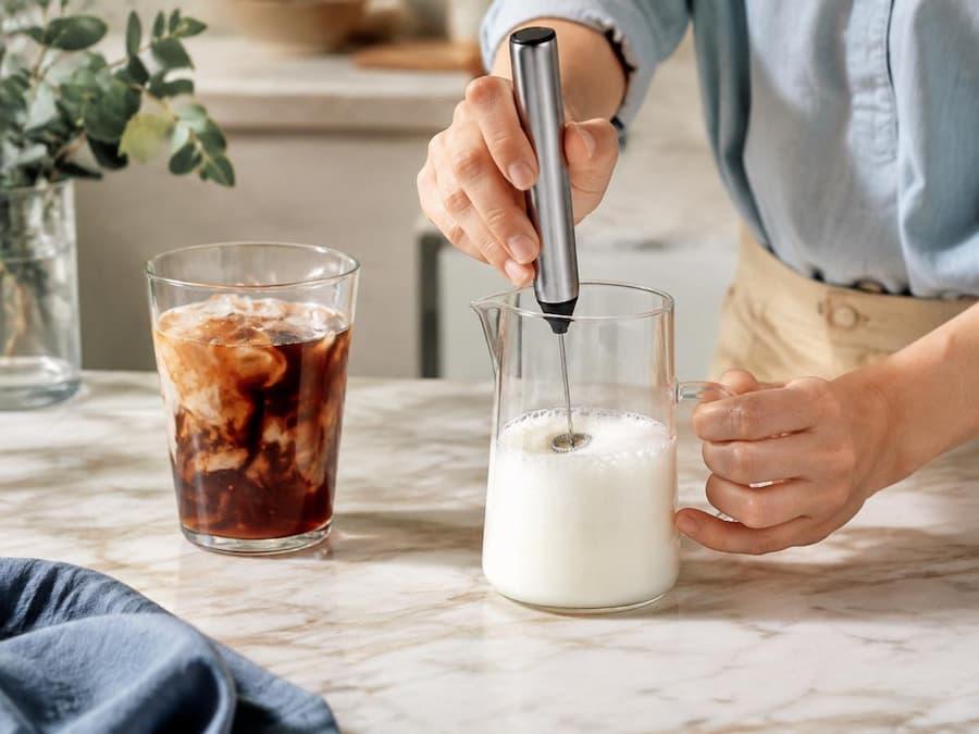 A handheld frother aerating cold milk beside a glass of iced coffee