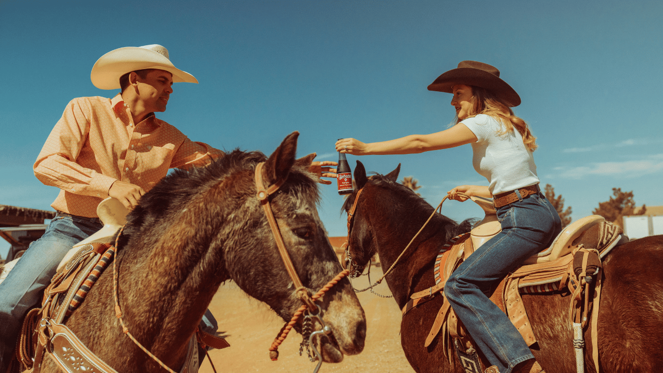 two people riding on a horse wearing a cowboy hat with drink