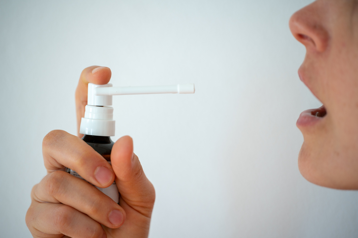 A profile image of a woman holding a sore throat spray bottle up to her open mouth. 