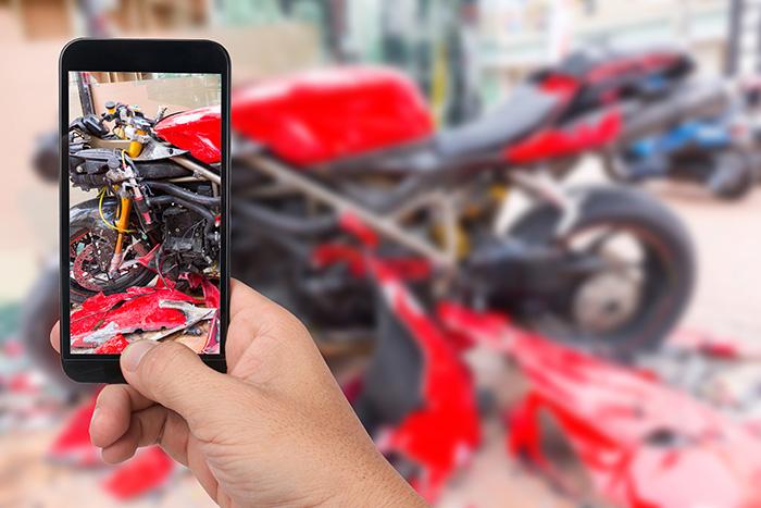 A motorcycle rider is using a smartphone to photograph the damage at an accident scene on a Texas highway, capturing crucial evidence for insurance claims and police reports. The rider may be documenting visible injuries and the condition of damaged vehicles to support their case with an experienced motorcycle accident attorney.