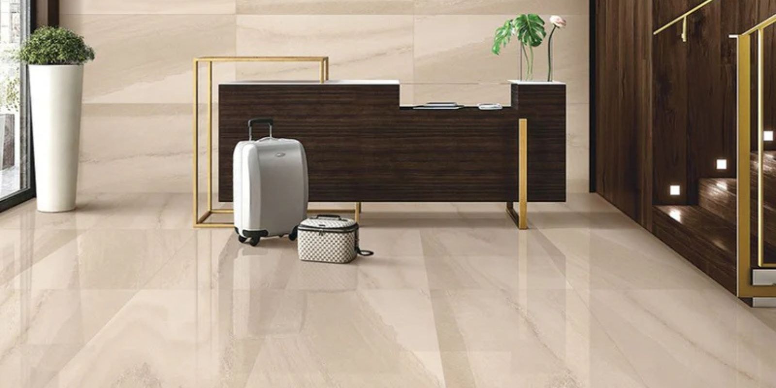 Modern hotel reception area with a wood and gold-accented desk, beige polished tile floor, luggage in front, and stairs to the right.