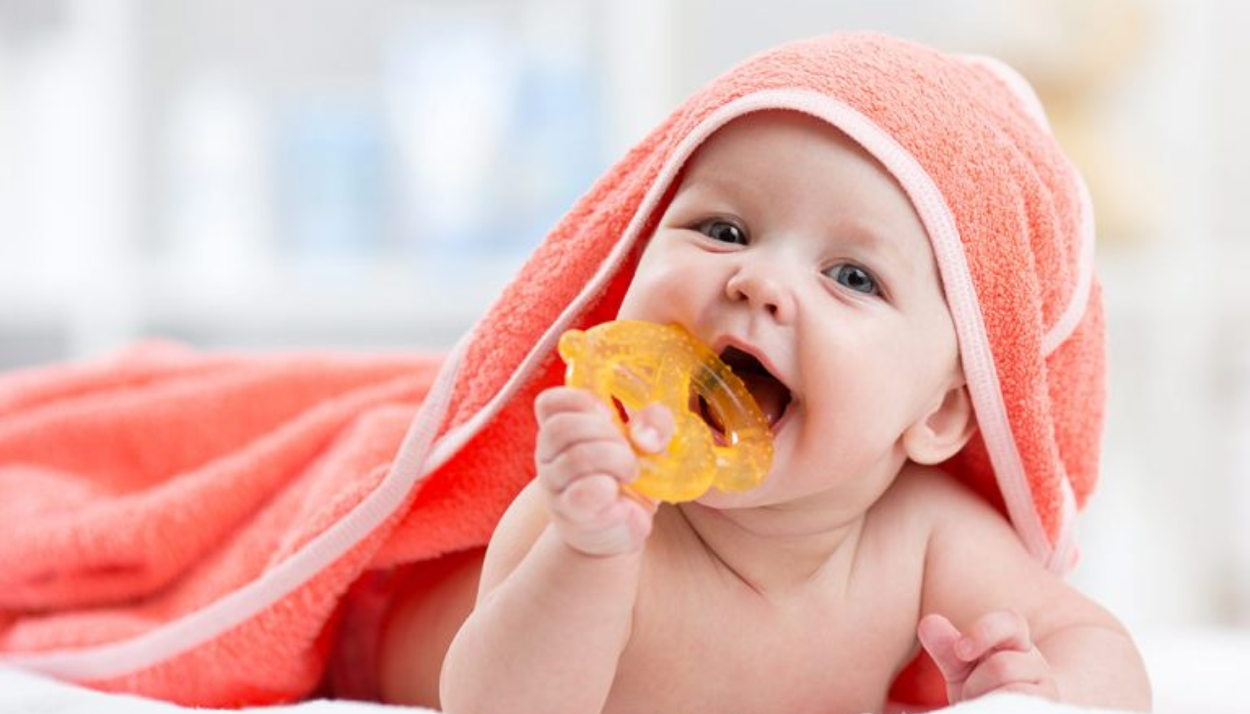 Baby ligger på magen med håndkle over hodet og biter på gul bitering.