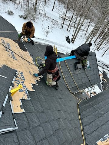 A picture of roofers installing a new roof in the winter. In the background, the trees are bare and the ground is covered with snow.