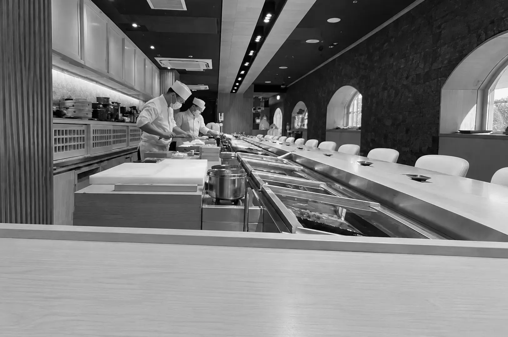 Two sushi chefs in white uniforms and hats prepare dishes behind a long counter in an elegant Japanese restaurant.
