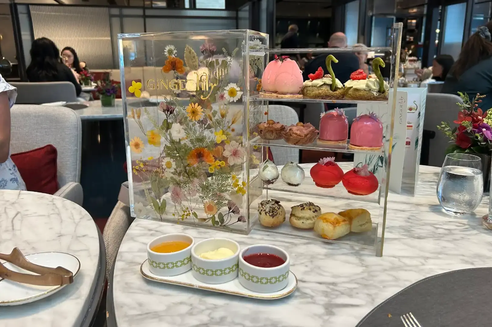 A white marble tabletop features a clear acrylic afternoon tea display stand adorned with pressed flowers on one side and multi-tiered shelves filled with colorful mini pastries, choux buns, and scones on the other. In front of the stand, three small ceramic ramekins containing honey, clotted cream, and berry jam rest on a matching oval tray.