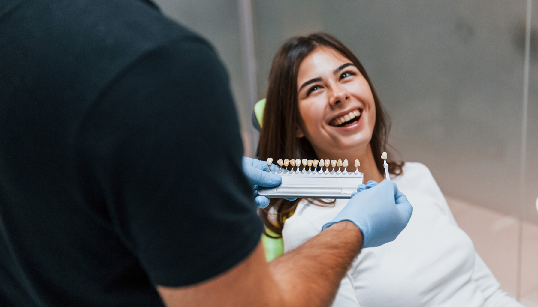 Tannimplantat i Norge: Hva Du Bør Vite Før Du Velger 18 Tannlege viser fargeskala for tannbleking til smilende kvinnelig pasient under konsultasjon.