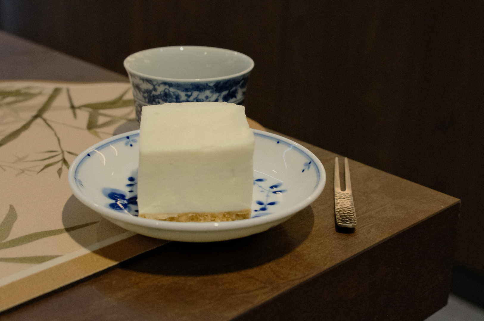 A square piece of creamy dessert on a white and blue plate beside a blue patterned cup. A textured fork lies on a wooden table, creating a serene, elegant vibe.