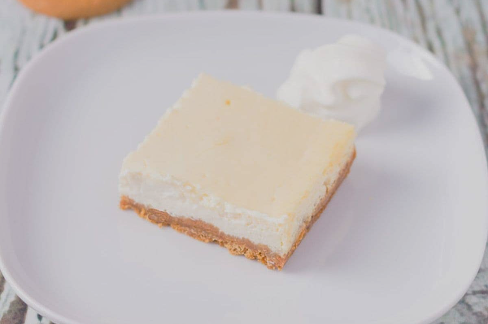A square cheesecake slice with a golden brown crust sits on a white plate. A swirl of whipped cream is beside it, creating an inviting, delicious appearance.