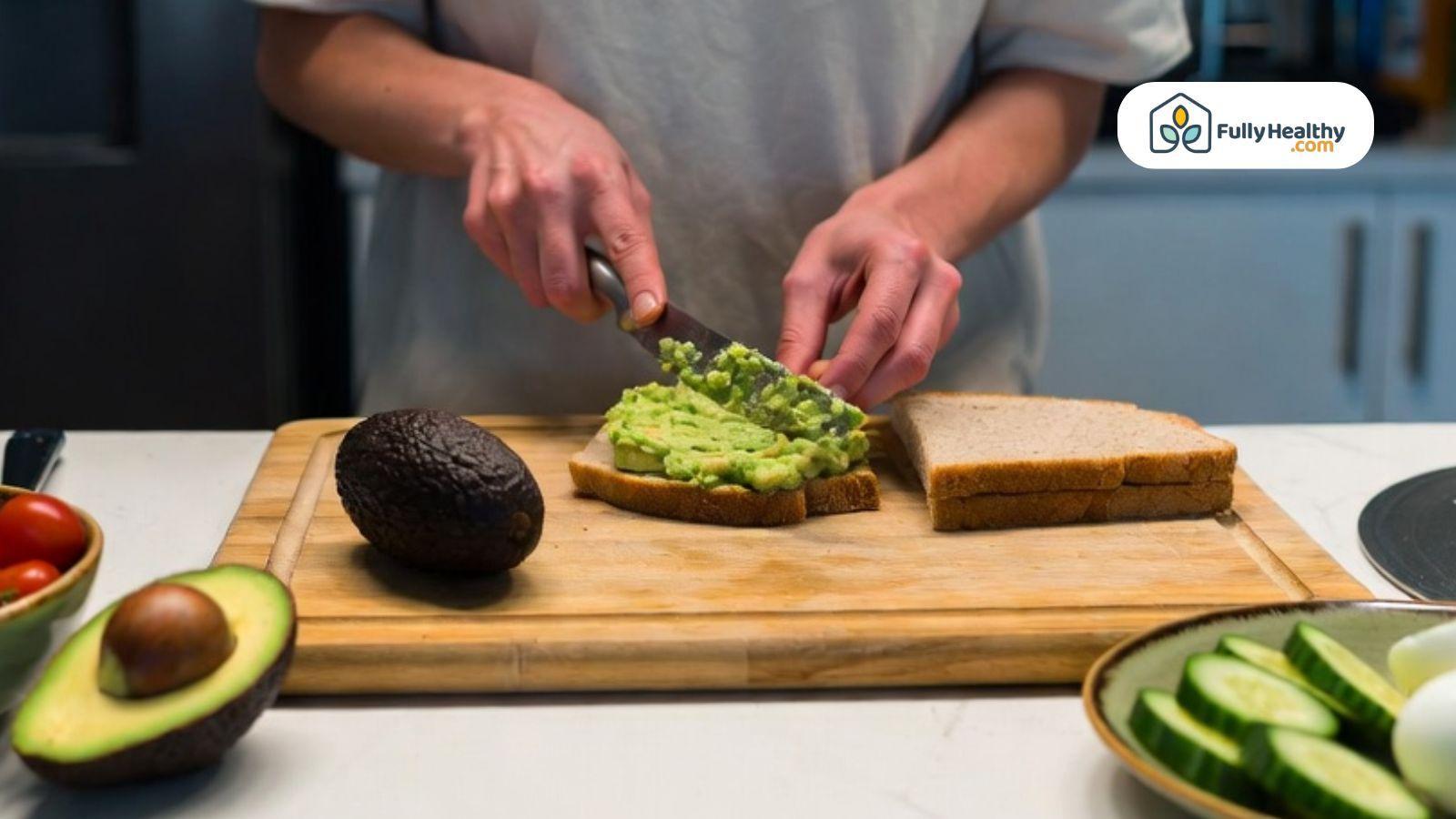 Person preparing mashed avocado toast with fresh vegetables in kitchen.