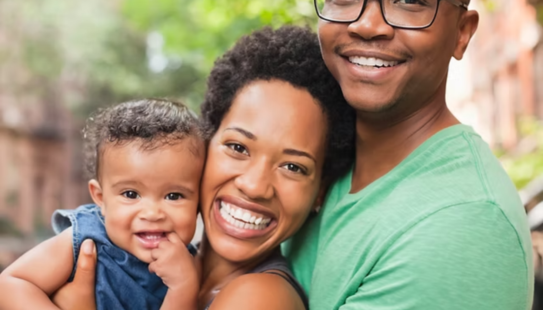 Smilende foreldre holder babyen mellom seg utendørs, alle ser glade og lykkelige inn i kameraet.