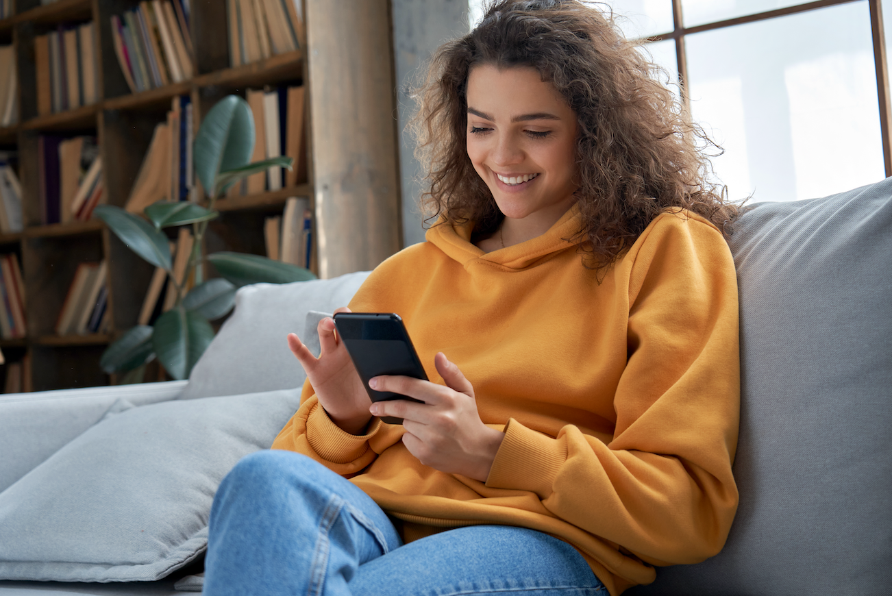 a woman in a yellow sweater and jeans smiles as she browses through her phone on the living room couch
