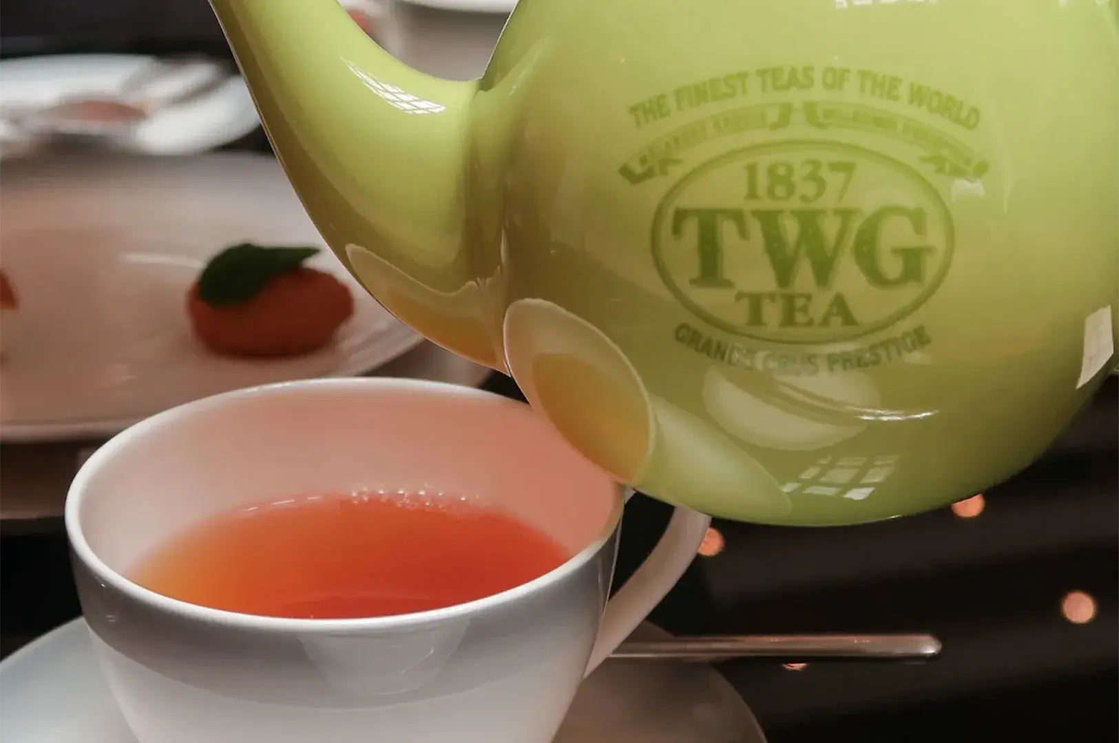 Close-up of a light green TWG teapot pouring fresh herbal tea into a white ceramic cup with a dessert blurred in the background.