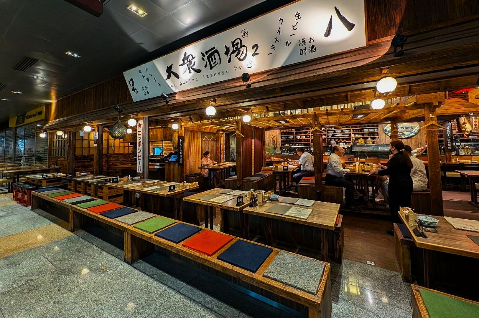 Cozy Japanese izakaya with wooden décor, colorful cushioned seating, and warm lighting. Customers enjoy meals, creating a welcoming atmosphere.