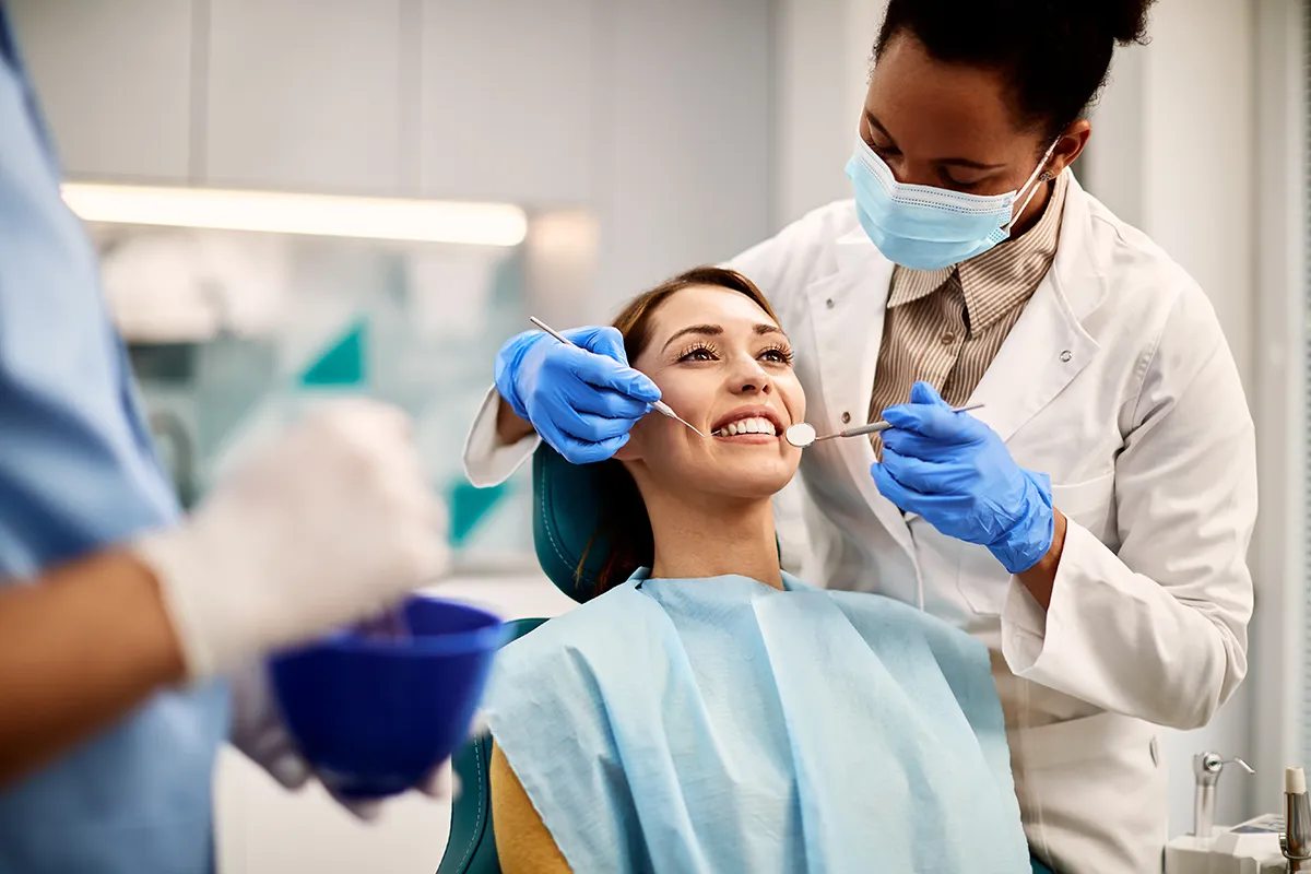 A dentist in a white coat and mask examining a female patient who is smiling in a dental chair.