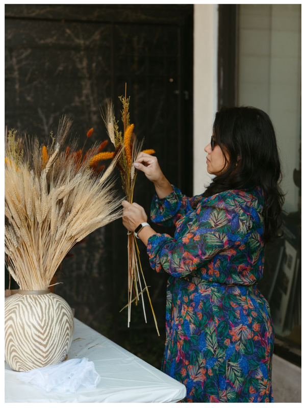 A woman arranges a fall-inspired floral display with dried wheat and orange stems in a neutral vase on a white table