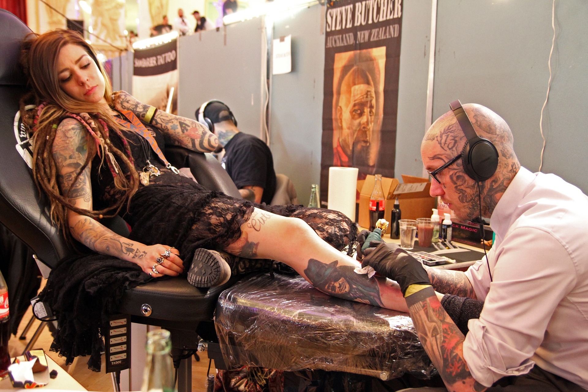Mulher ruiva, com longos cabelos lisos e pele clara, usando roupa preta, com tatuagens nos braços e nas pernas, está deitada sobre a cadeira de um tatuador em um estúdio, enquanto o tatuador faz uma nova tatuagem em sua perna. O tatuador tem a pele clara e a cabeça raspada, com tatuagens no braço e na cabeça. Imagem: Licel, Pixabay.com.  
