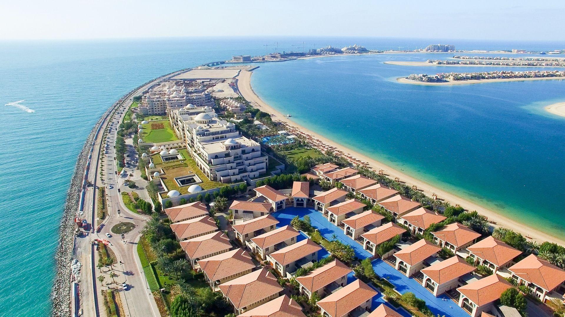 Aerial view of a luxury beachfront residential community on a palm-shaped island, featuring rows of villas with terracotta roofs, landscaped streets, private pools, and a long sandy shoreline along clear blue water.