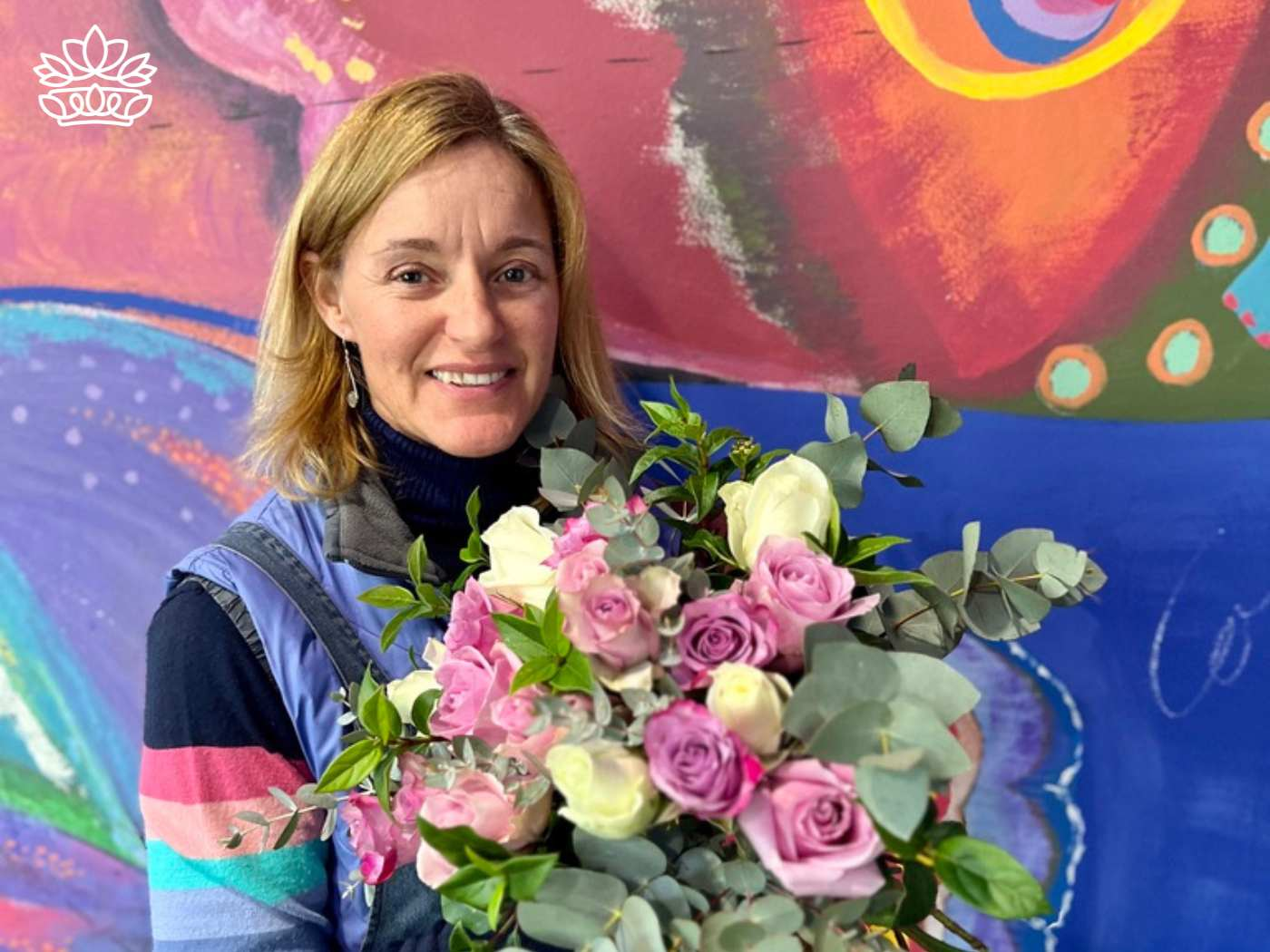 A florist with a warm smile, holding a delicate flower bouquet of pink and white roses interspersed with silvery eucalyptus, standing in front of a colorful mural, representing the Flowers By Occasion Collection at Fabulous Flowers and Gifts.